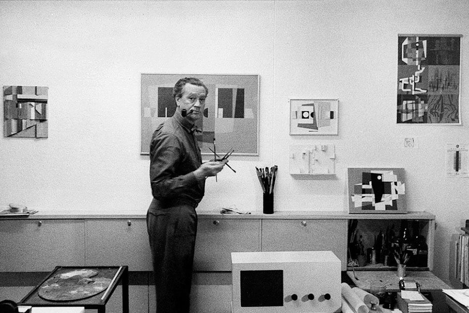 Robert S. Gessner in seinem Atelier in Z&uuml;rich, 1958. Der K&uuml;nstler pr&auml;sentiert sich mit Pinseln in der Rechten und der Pfeife im Mund. (Bild: Michael Wolgensinger / Archiv RSG / &copy; Archiv Fotoatelier Wolgensinger)