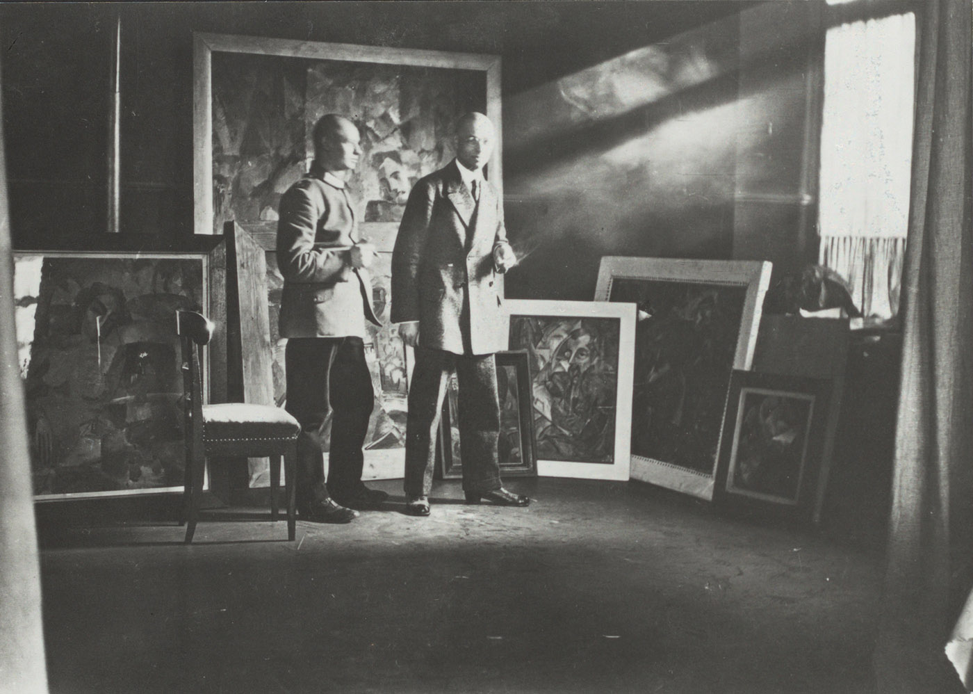Johannes Itten in seinem Atelier zusammen mit Oskar Schlemmer, Stuttgart 1913, im Bildhintergrund seine frühen abstrakten Gemälde (Hs NL 11: Ba 3.7; <a href="https://doi.org/10.7891/e-manuscripta-123165">Digitalisat</a>)